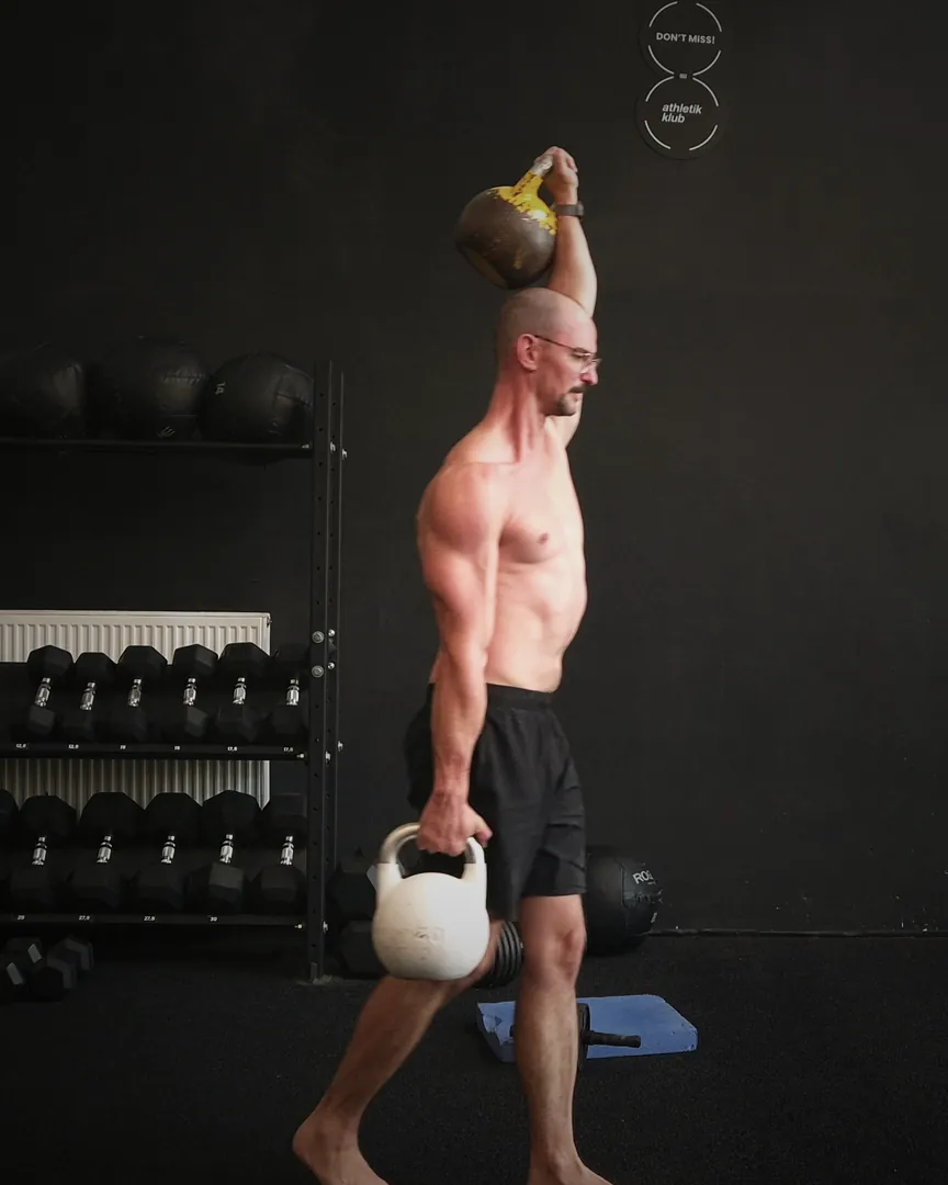 Bene trägt eine Kettlebell über dem Kopf und eine an der Seite